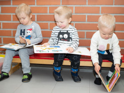 Kolme pientä lasta istuu penkillä tiiliseinän edessä ja katselee kuvakirjoja. 