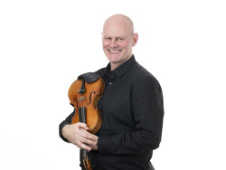 Tummapaitainen kalju mies pitää sylissään viulua. Kuva Jiri Halttunen
