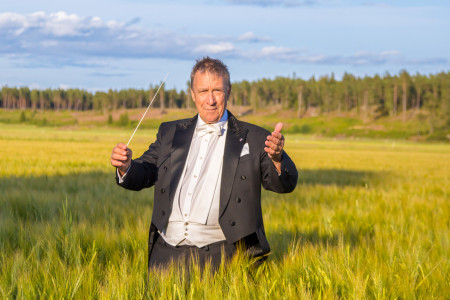 Frakkiin pukeutunut mies seisoo pellolla viljan keskellä ja taustalla näkyy sininen taivast sekä metsää. Kuva Jyväskylä Sinfonia